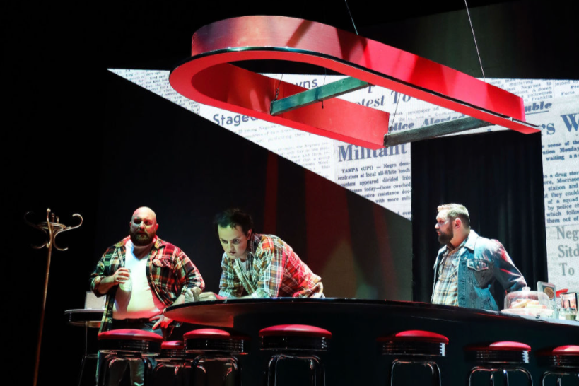 Three men in plaid shirts standing around a bar with red stools in a dimly lit space, with a large red and black circular decoration hanging overhead.