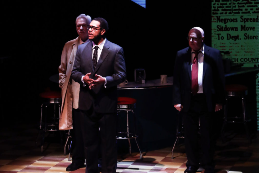 Three men in suits standing on a stage with a dark background and bar stools, one man speaking and two listening.
