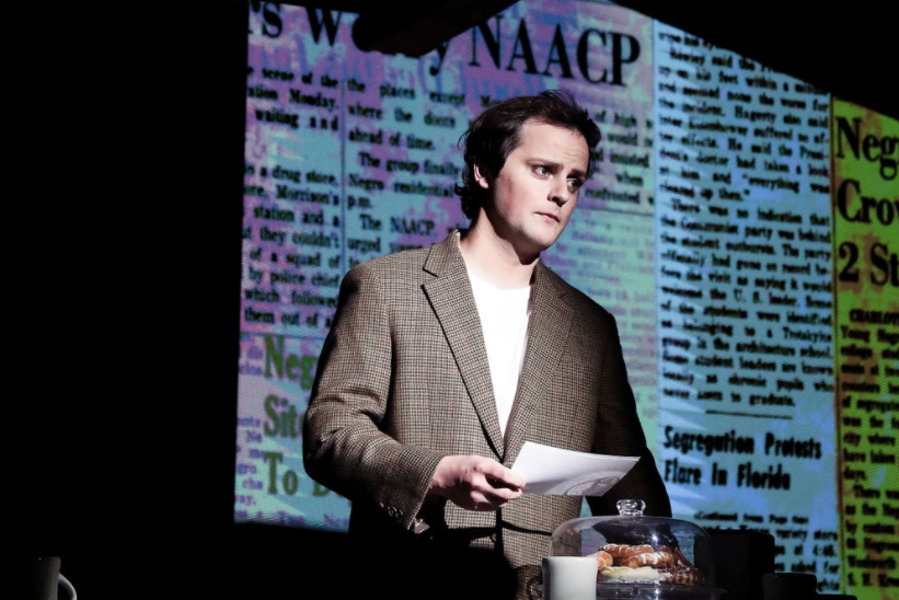A person standing on stage in front of projected newspaper headlines, wearing a brown blazer and holding a piece of paper.