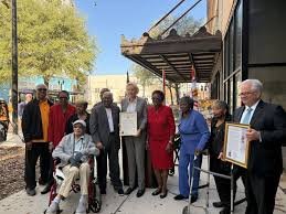 A group of people gathered outside a building for a photo, with one person holding a framed certificate and another holding a sign.