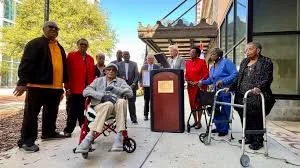 Group of people, including an elderly man in a wheelchair, standing and sitting outside near a building, some with walkers and others with bags, on a sunny day.