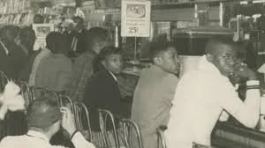 A black and white photograph of a busy diner with several people sitting at the counter and tables, some engaged in conversation.