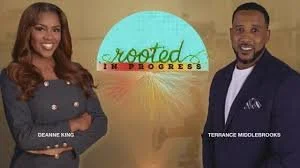 Two people standing in front of a backdrop with the words 'Rooted in Progress' and a sun design. The woman on the left has long hair and is smiling, wearing a dark top. The man on the right is wearing a suit and tie, also smiling.