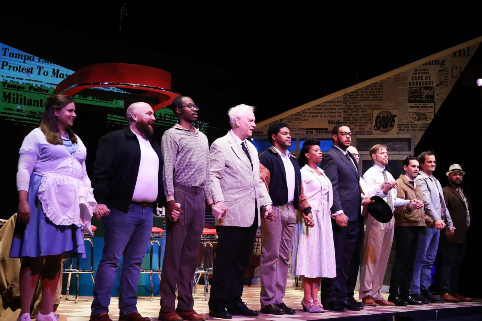 A group of people holding hands on a stage, possibly at a performance or ceremony, with a background featuring posters and newspapers.