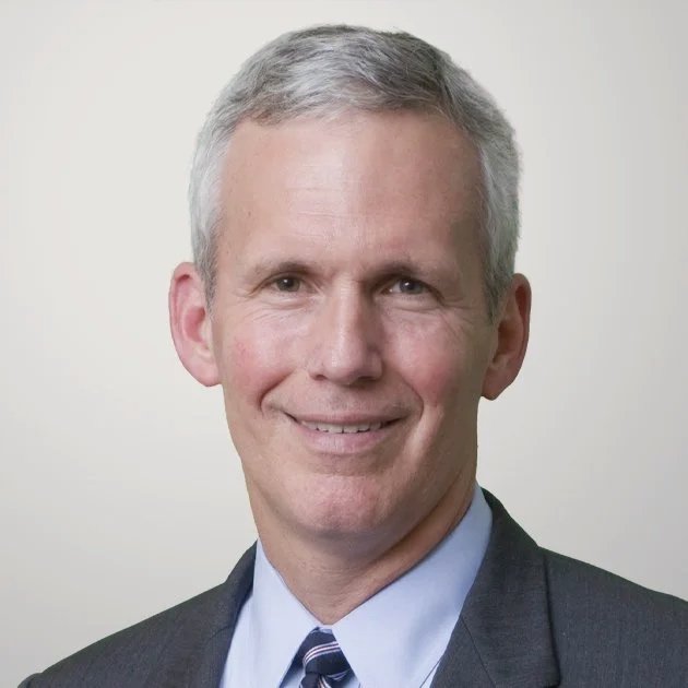 Headshot of a middle-aged man with short gray hair, wearing a dark suit, light blue dress shirt, and striped tie, smiling at the camera against a plain light background.