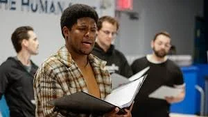 A man singing or speaking passionately during a rehearsal or practice, with three other people holding scripts or papers in the background.