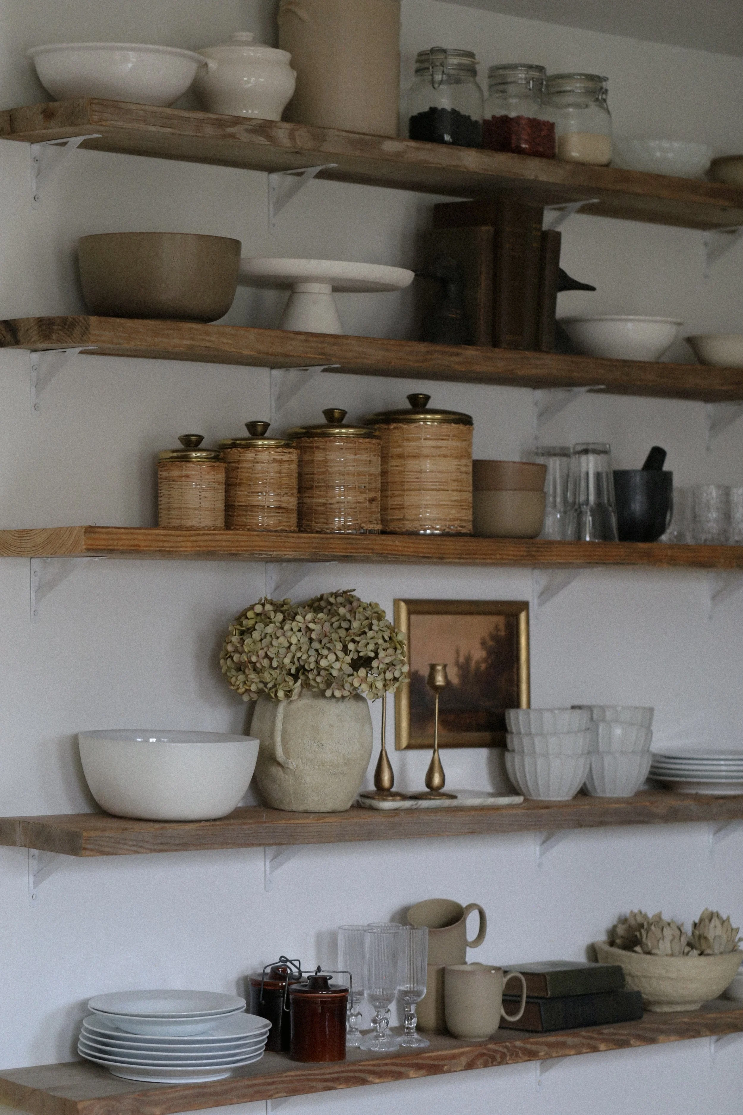 Kitchen open shelves with rattan canisters, dinnerware, and pitcher with hydrangeas. Cottage core, earthy, antique