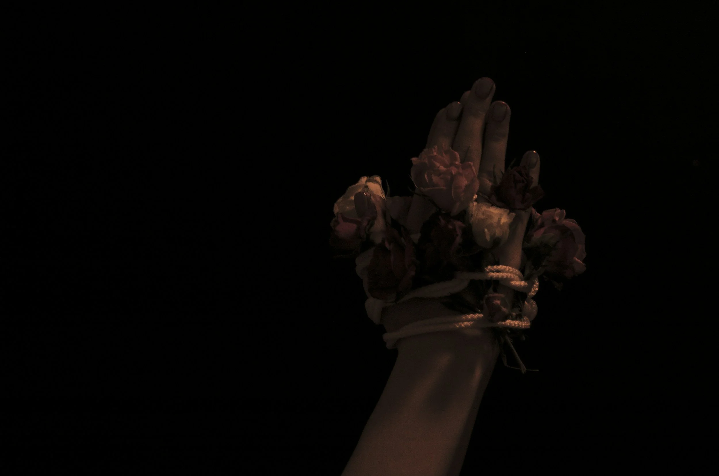 A hand raised vertically with fingers extended against a black background, adorned with multiple pink and cream-colored flowers and white cord wrapping around the wrist.