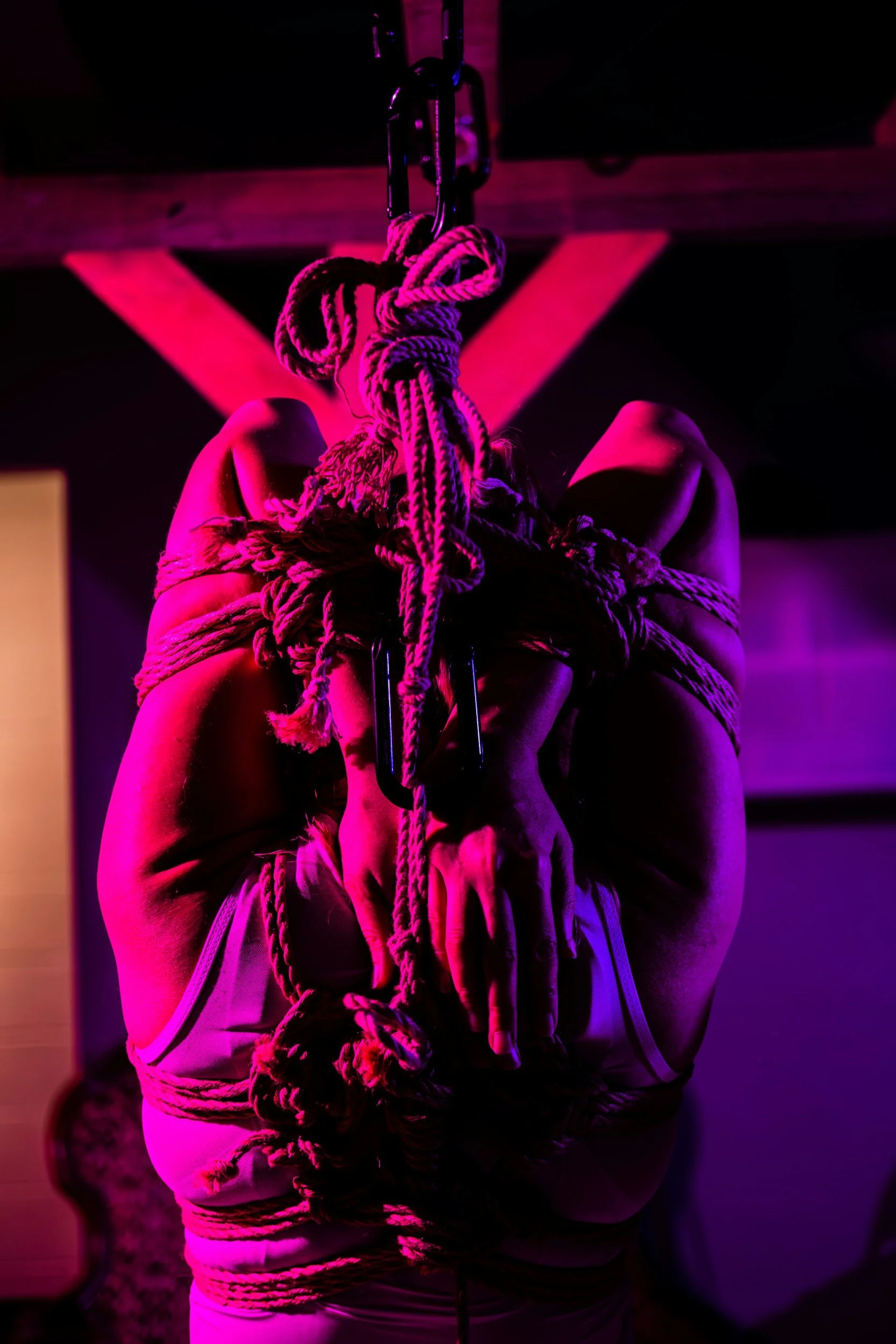 A person hanging upside down, bound with ropes, suspended from a hook in a dark room illuminated with purple and pink lighting.