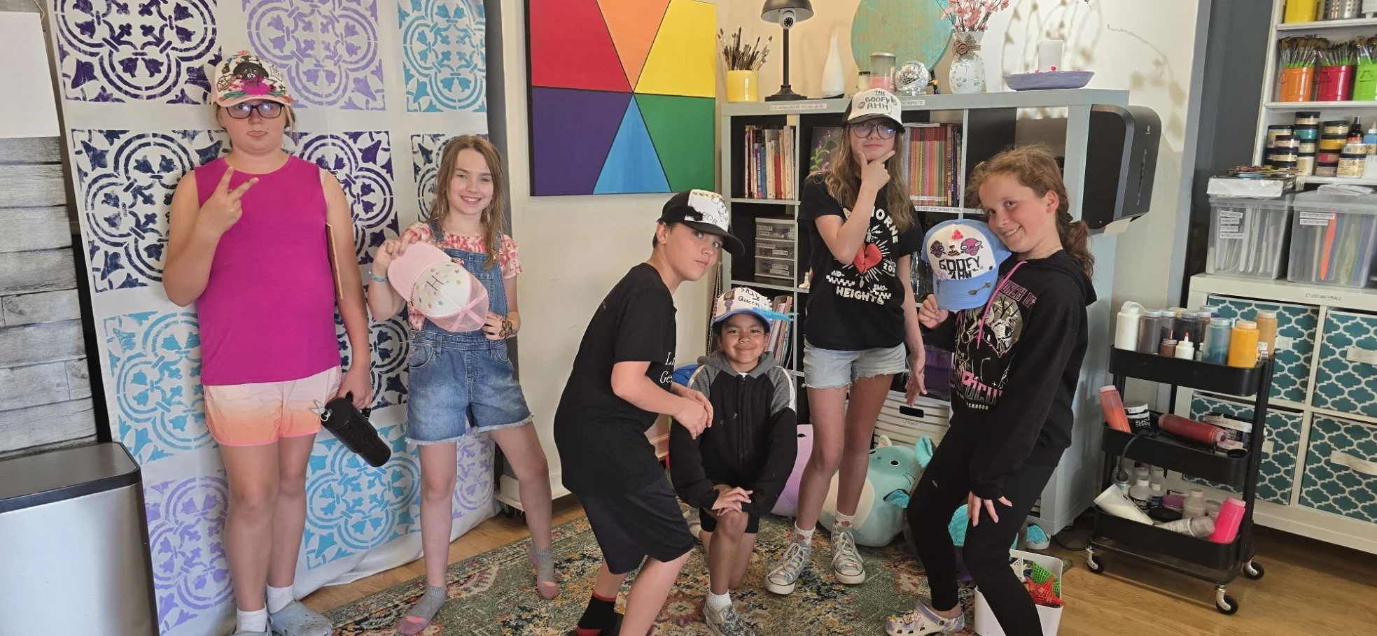 Group of seven children posing indoors in a room with colorful decor, some wearing hats, and holding various craft items. There is a vibrant geometric painting on the wall, shelves with art supplies, and a patterned backdrop.
