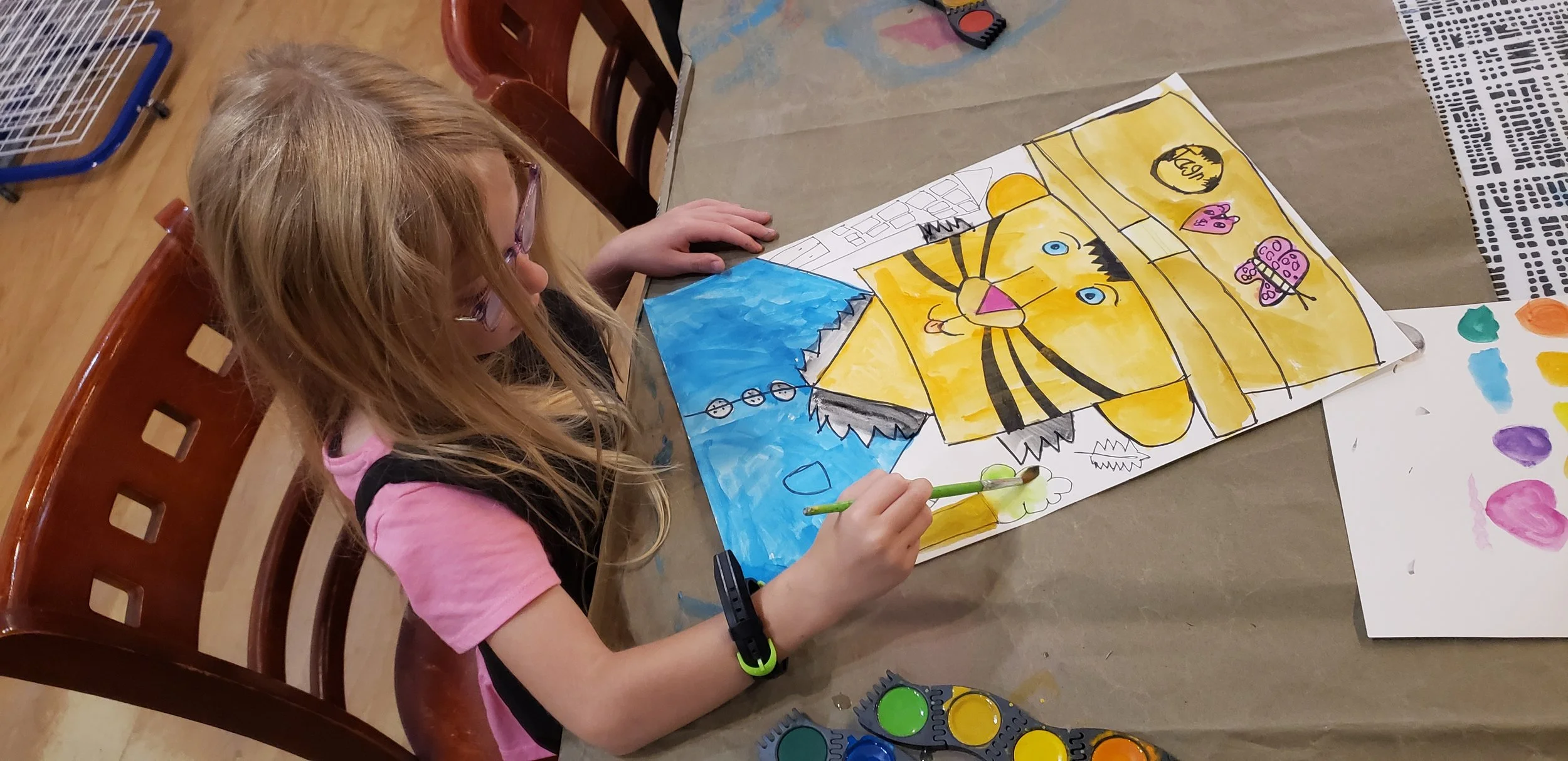 A young girl with glasses and a pink t-shirt painting a colorful poster of a yellow tiger with black stripes, blue eyes, and a pink nose, surrounded by various painted shapes and cartoon characters on a table.
