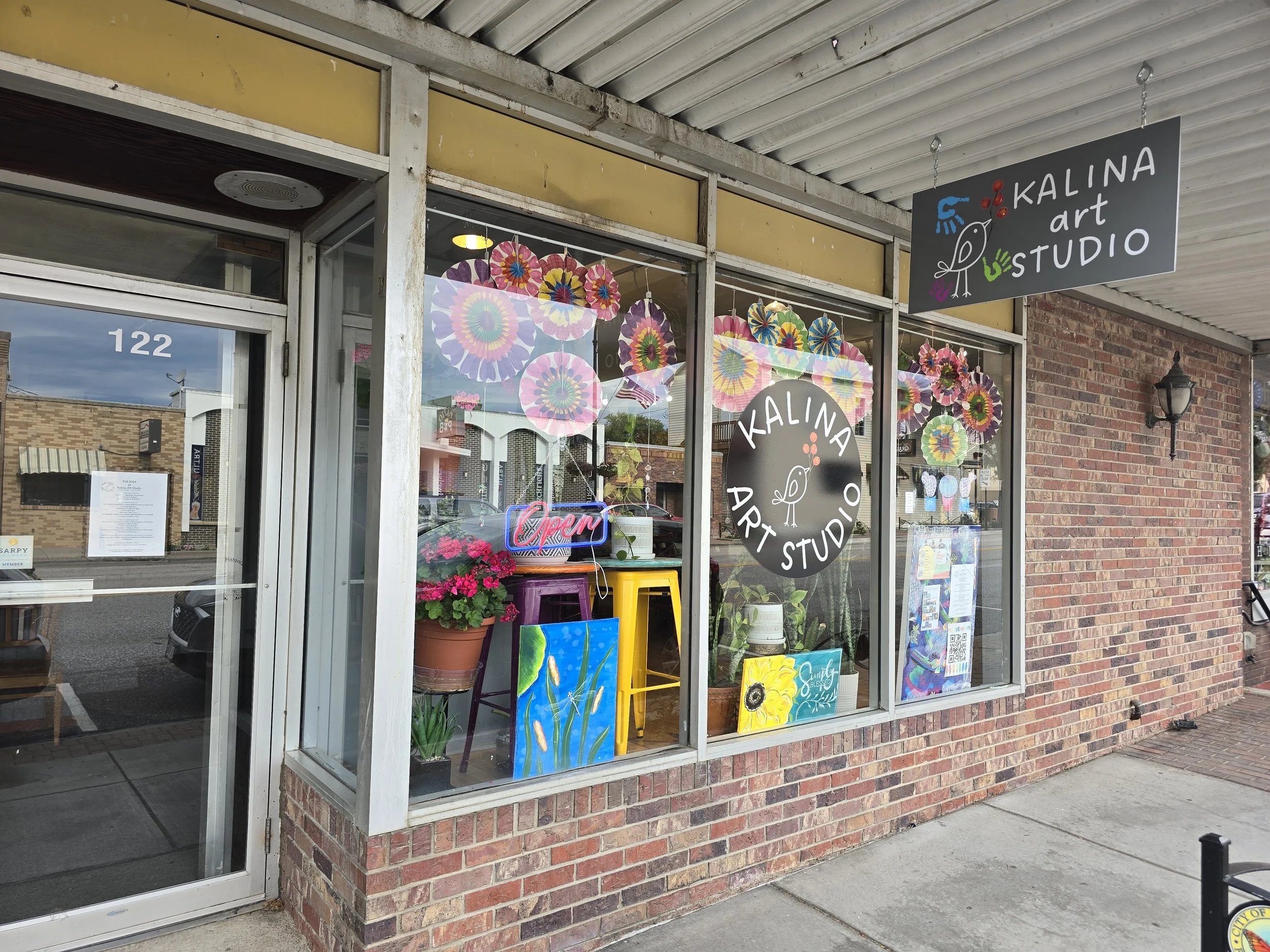 Exterior view of Kalina Art Studio storefront with colorful paper pinwheels, flowers, paintings, and a neon 'Open' sign in the window.