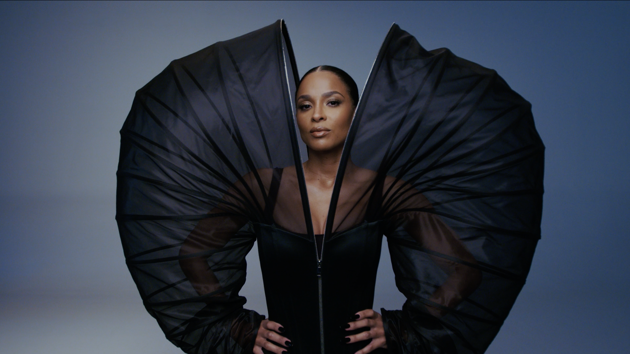 A woman with dark skin and sleek hair poses confidently, wearing a black dress with large, dramatic black fabric wings or structures extending from her shoulders, set against a gradient gray background.