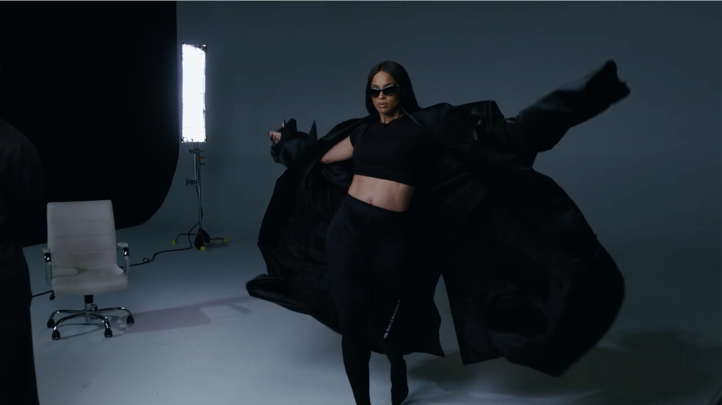 A woman in black clothing and sunglasses, posing in a photography studio with a dark gray background, Studio lights, and a white chair.