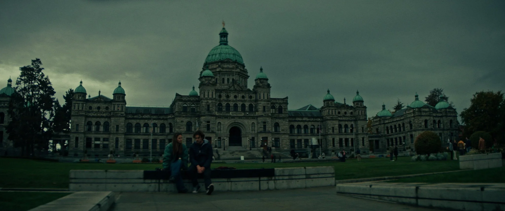 A large historic government building with domes on its roof, set against a cloudy sky. Two people are sitting on a bench in the foreground near a well-kept lawn.