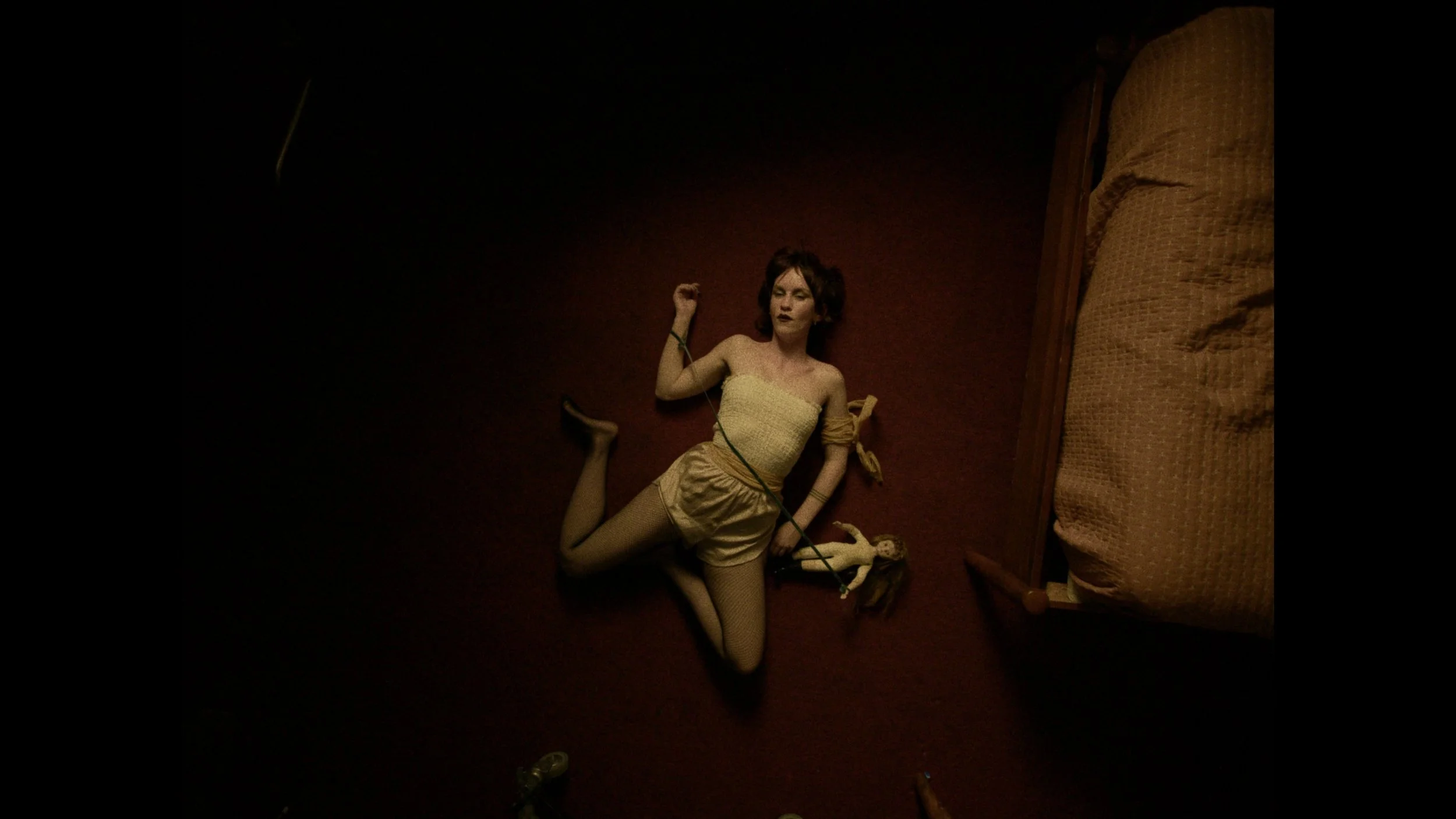 A woman lying on a dark carpeted floor next to a bed. She is wearing a beige strapless top, shorts, and fishnet stockings. Her eyes are closed, and her hair is brown and styled in soft curls. She appears to be holding a long black object, possibly a 