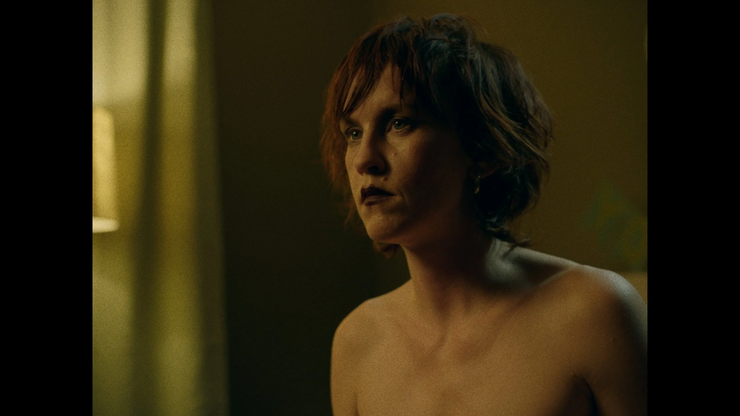 A woman with short, tousled brown hair sits topless in a dimly lit room, gazing thoughtfully to the side with a serious expression.