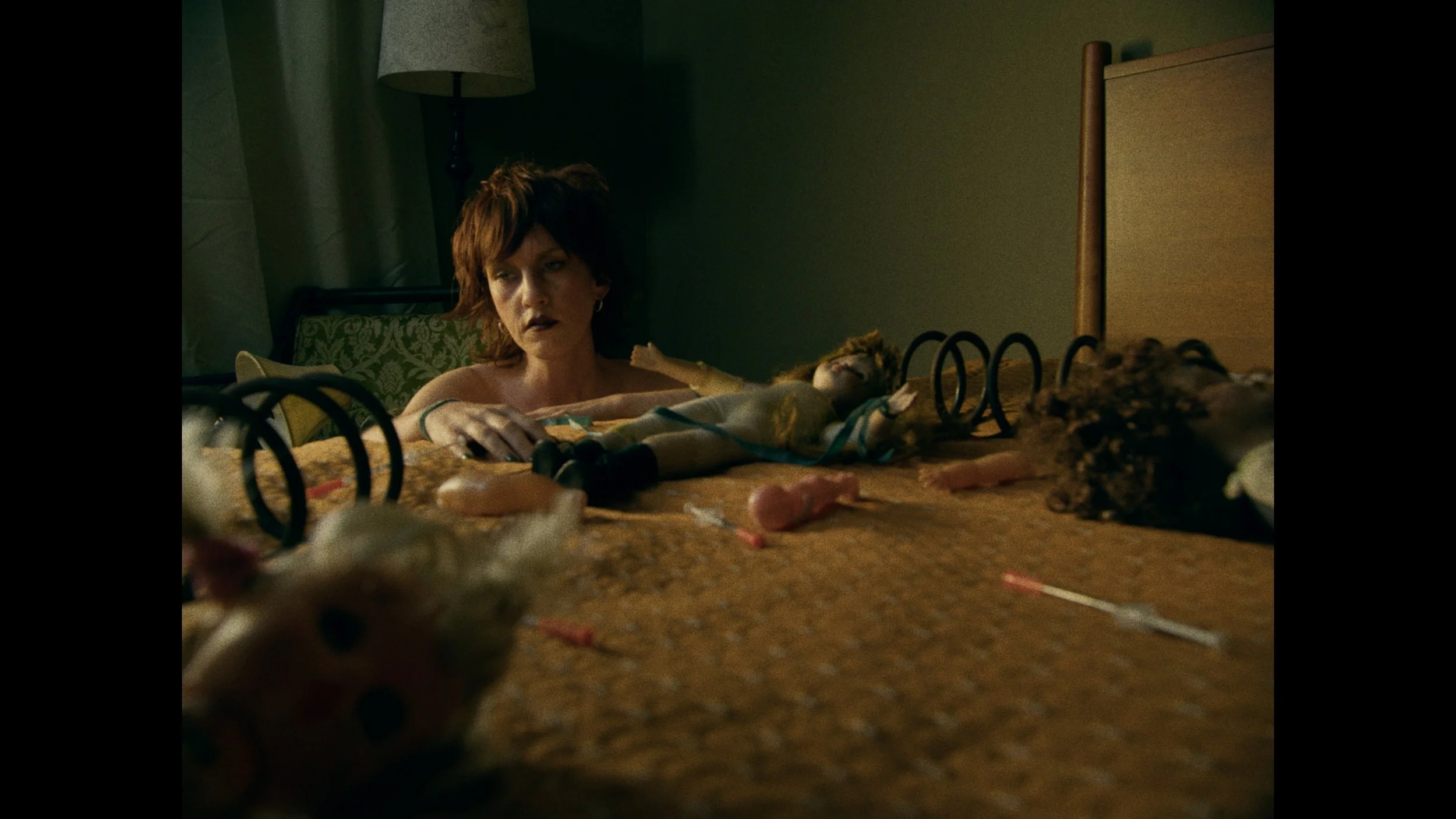 Woman with short brown hair lying on a bed surrounded by toys and dolls in dim lighting.