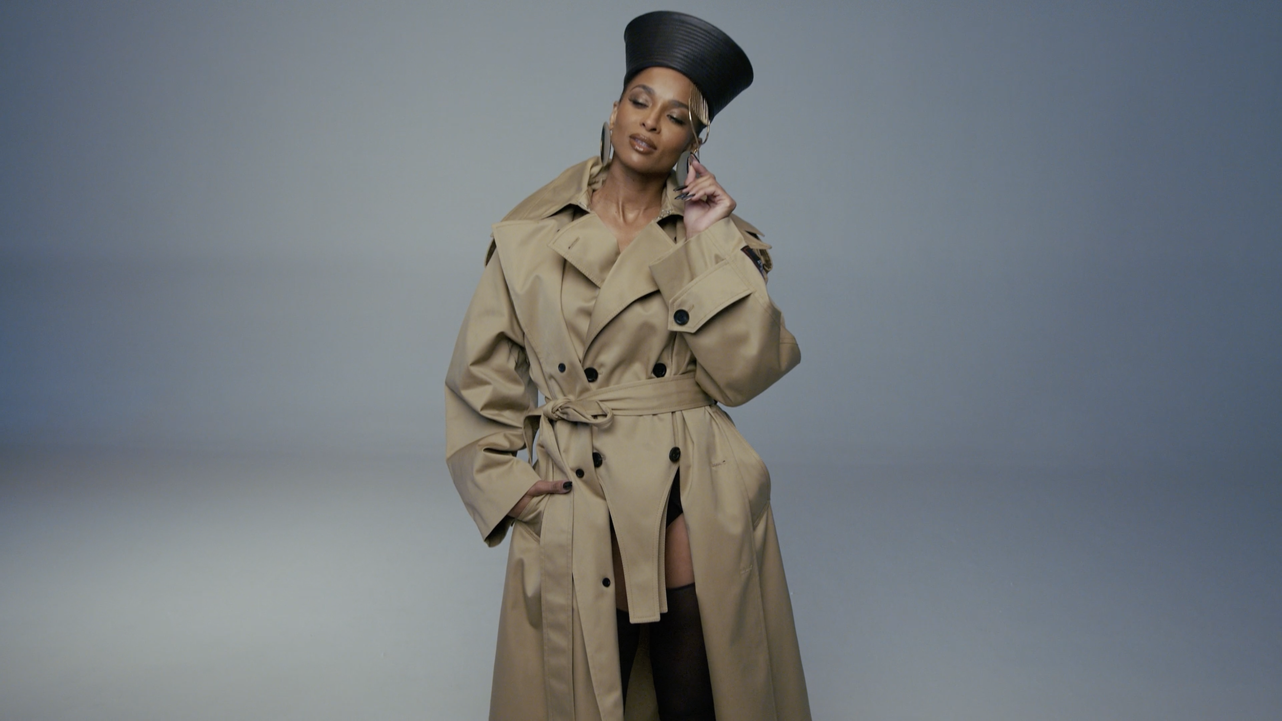 A woman wearing a tan trench coat, black thigh-high stockings, and a large black hat, standing against a plain gray background. She is holding a phone to her ear with her left hand and has her right hand in her pocket.