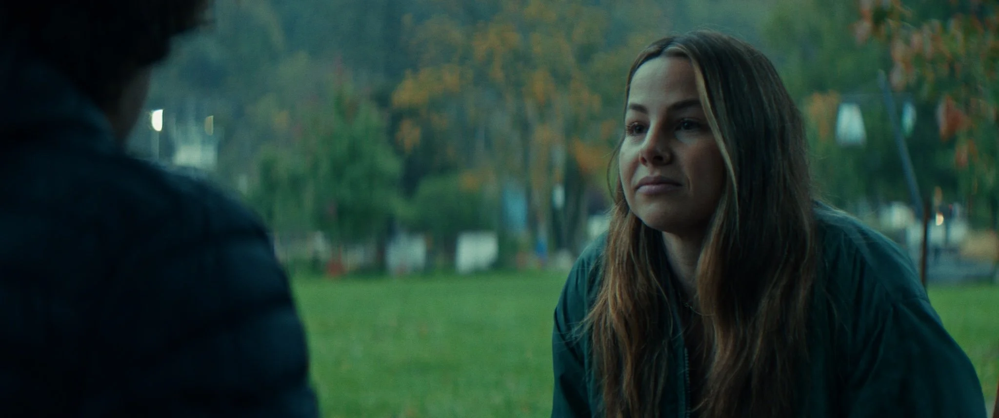 A woman with long brown hair and a serious expression sitting outdoors in a park during autumn, wearing a dark jacket.