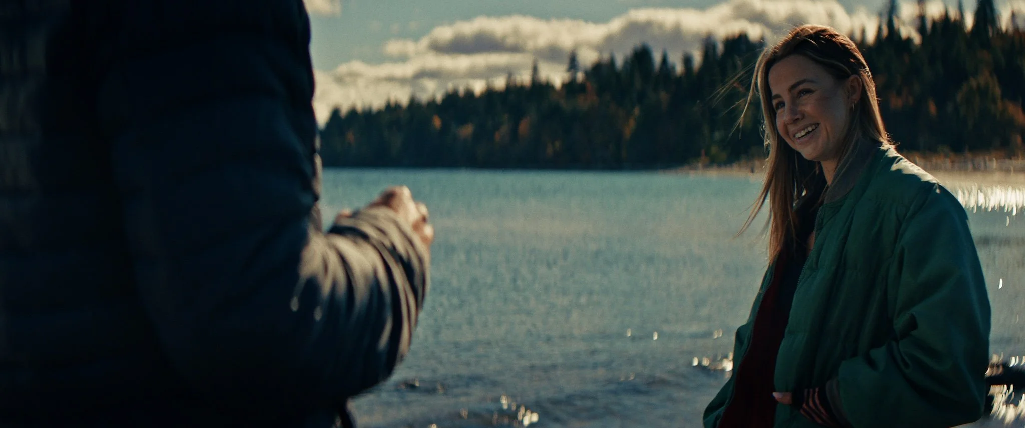 A woman with red hair wearing a green jacket smiling by a lake during daytime, with a person partially visible on the left.