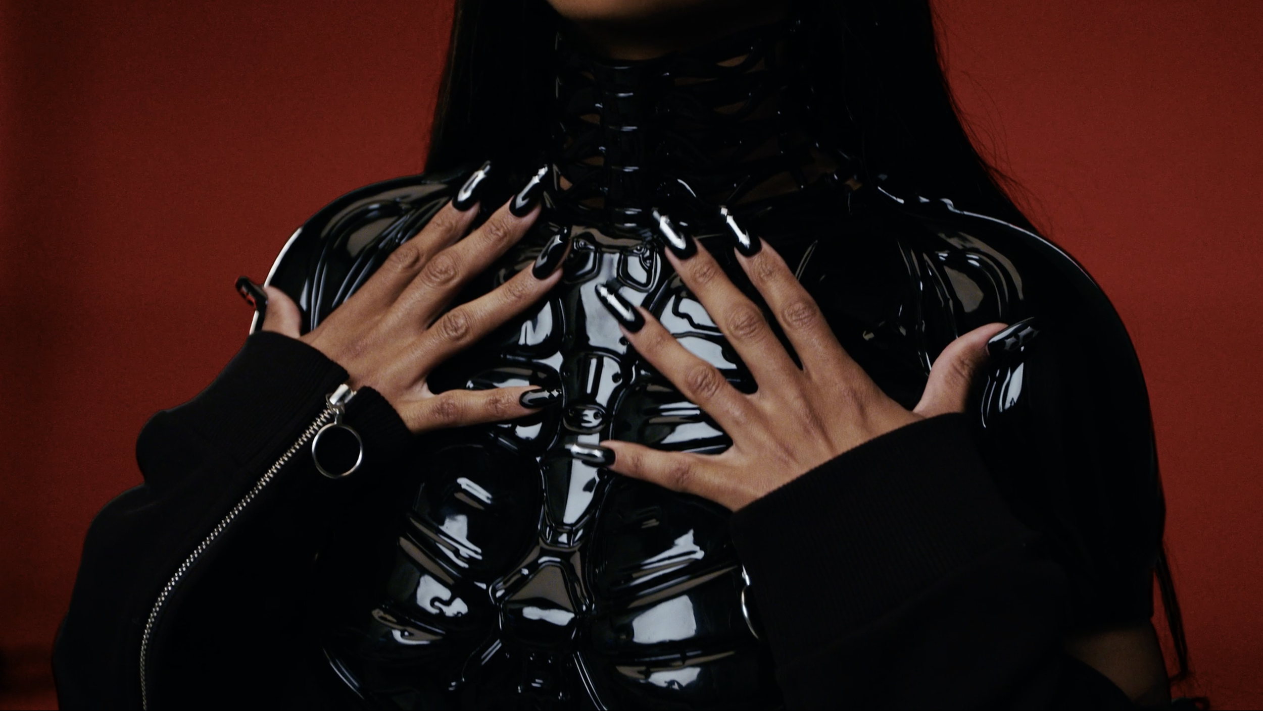 Person wearing a black, shiny, latex or PVC-looking top, with hands crossed over the chest, showing black long nails with white streaks and a ring on one finger. The background is dark red.