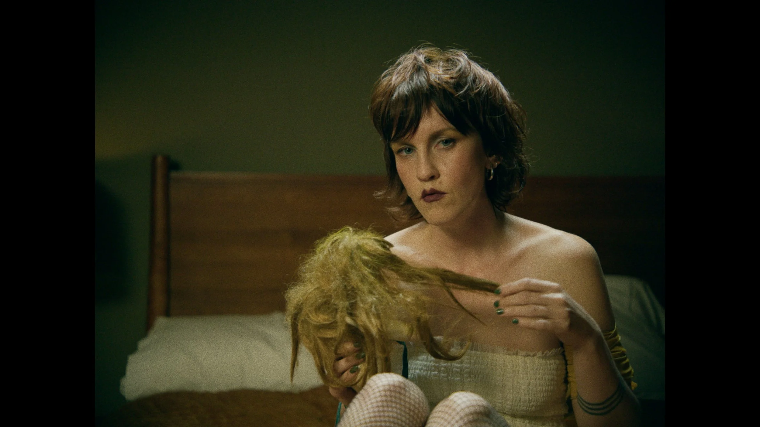 A woman with short brown hair and dark lipstick sitting on a bed, holding a doll with blond dreadlocks, in a dimly lit room with green walls.