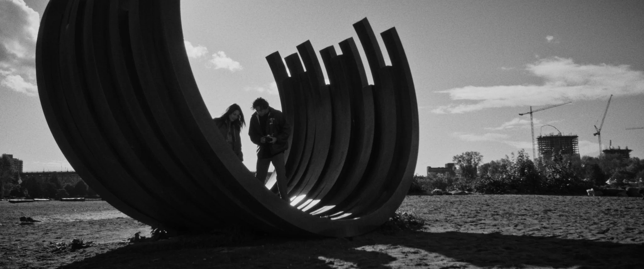 Silhouette of two people standing inside a large, curved, wooden sculpture on a sandy area with a cityscape, including construction cranes, in the background during daytime.