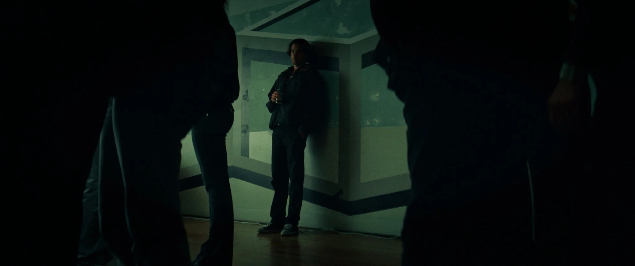 A person standing and holding a drink in a dimly lit room, surrounded by other people whose legs are visible in the foreground.