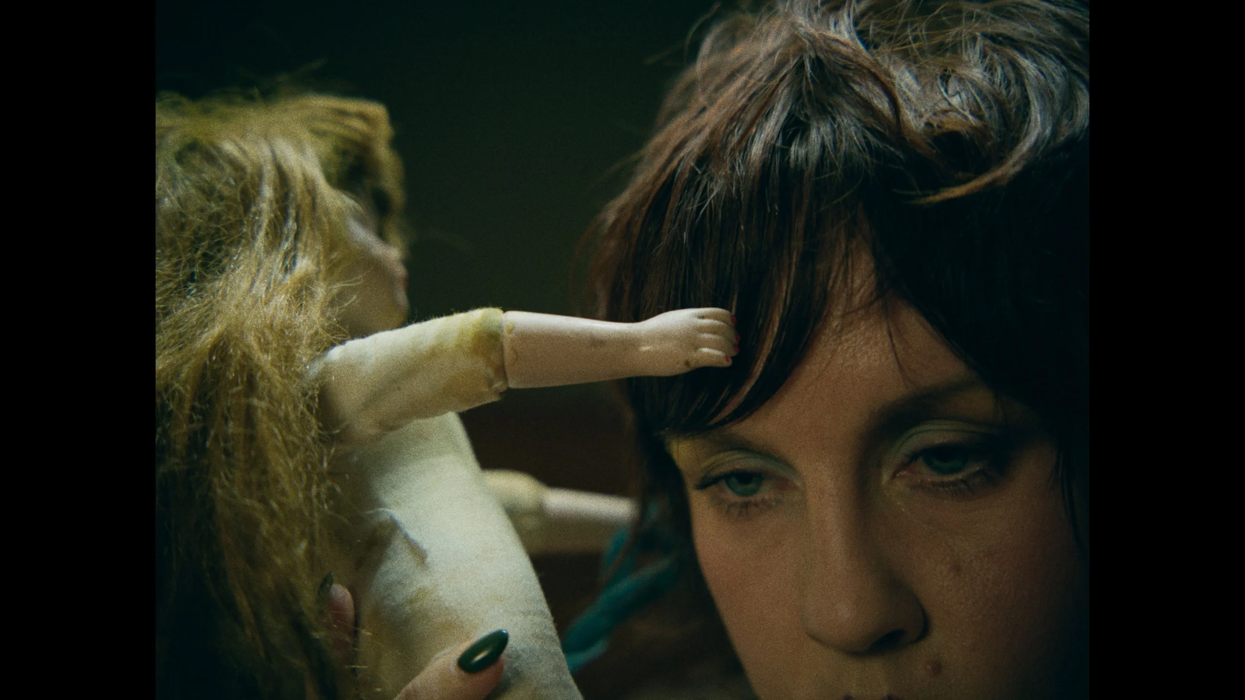 Close-up of a woman with short dark hair and green eyes, looking down, with a small doll's hand touching her head.