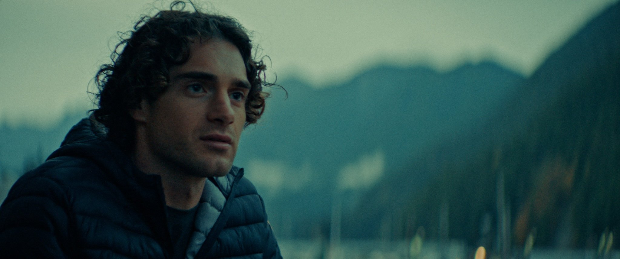 A man with curly hair and a black puffy jacket is sitting outdoors, with mountains and a lake visible in the background, during what appears to be dusk or dawn.