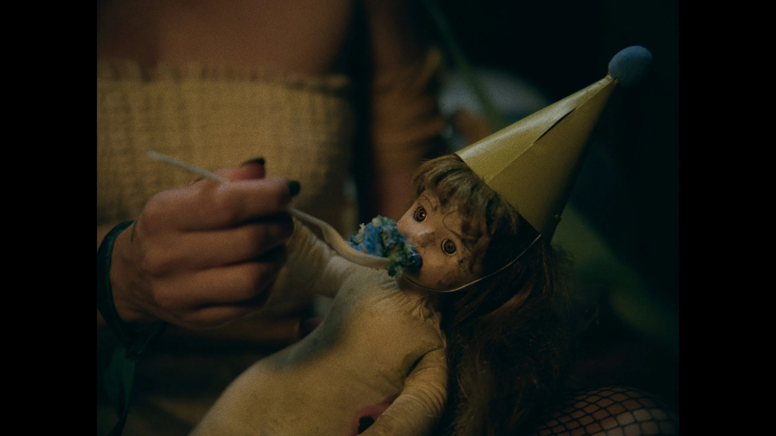 A person in a yellow top with a tattoo on their wrist is feeding a doll with a spoon. The doll has brown hair, big eyes, and is wearing a yellow dress and a gold party hat. The doll's mouth has blue frosting.