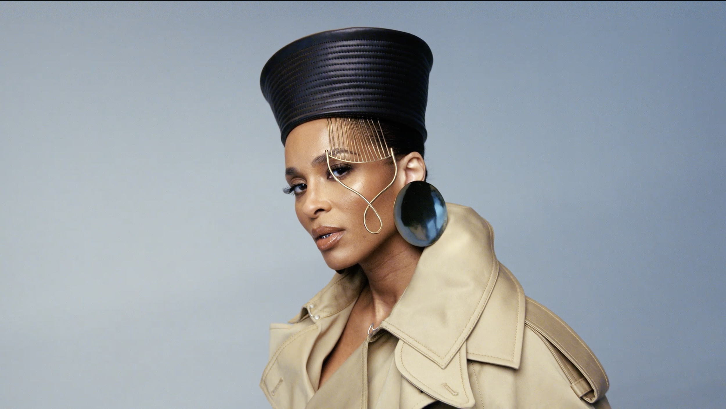 A woman wears a large black hat with gold decorative chains, large black circular earrings, a beige trench coat, and is posed with her head slightly turned, looking at the camera against a plain light background.