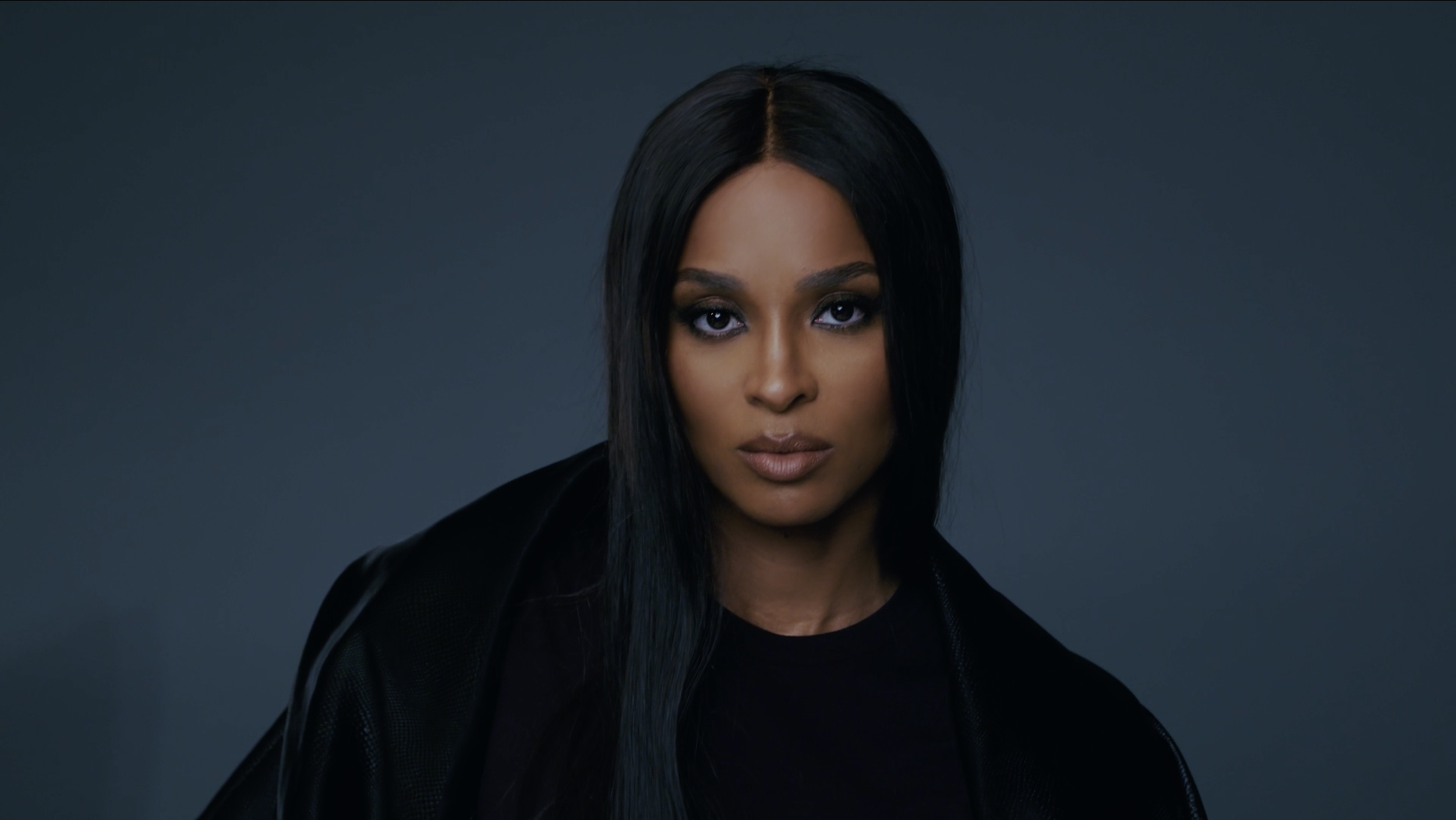 A woman with long black hair, wearing a black outfit, looking directly at the camera against a dark background.