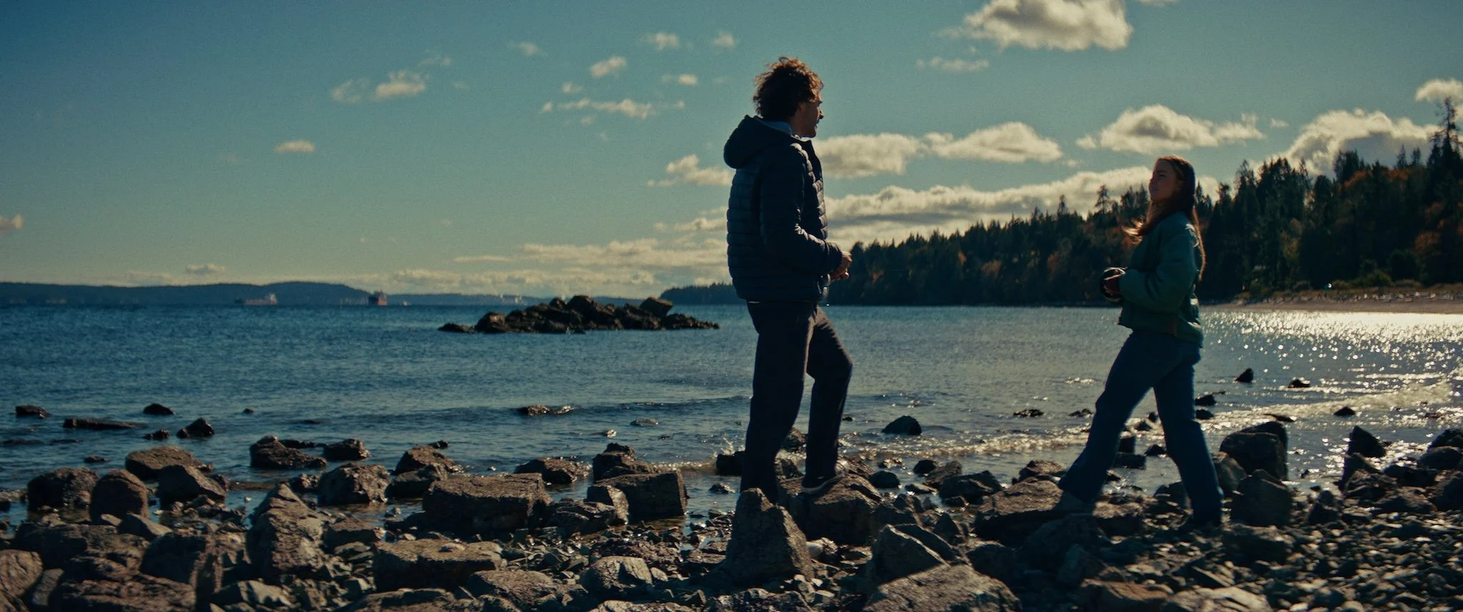 Two people, a man and a woman, stand on a rocky shoreline by the water, with trees and clouds in the background during daytime.