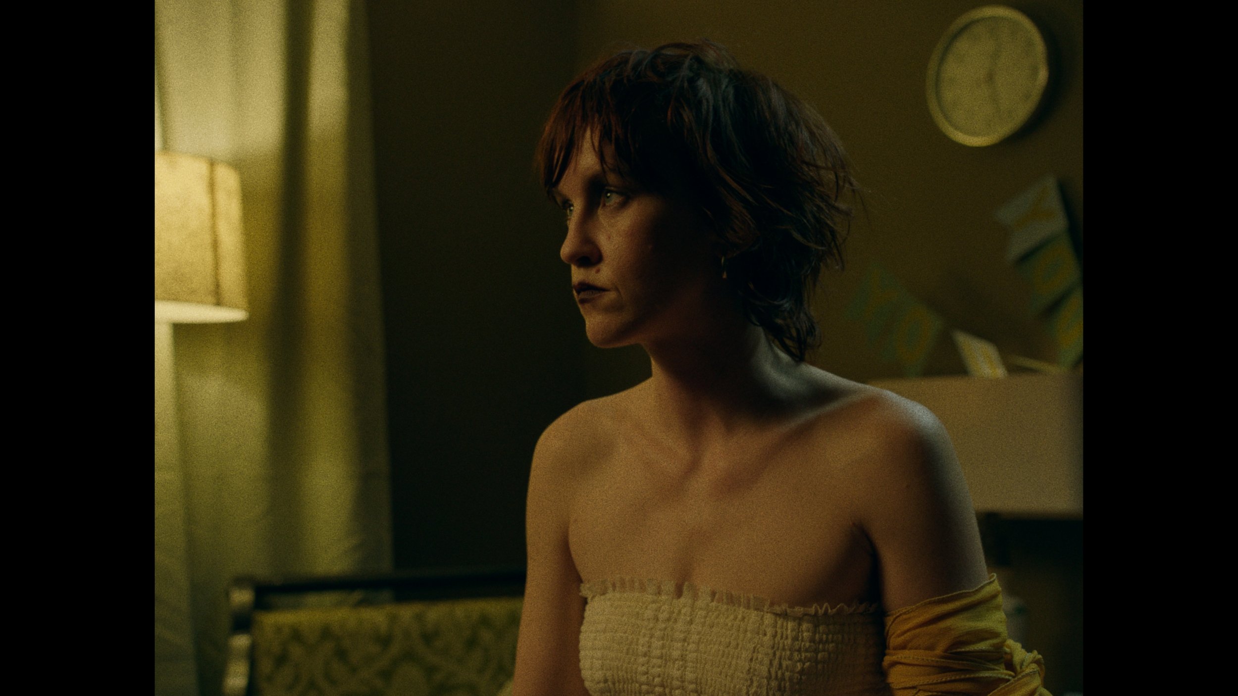 A woman with short, wavy dark hair looking to the left in a dimly lit room, wearing a strapless top and an arm covering over her shoulders.