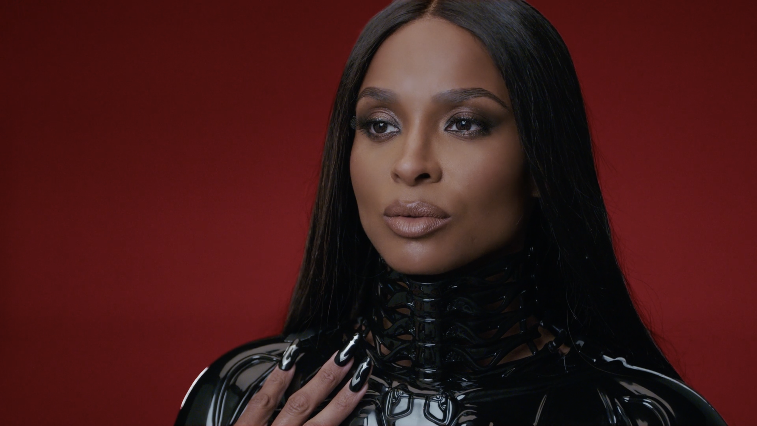 A woman with long dark hair and makeup, wearing a black latex outfit, touching her neck, against a red background.