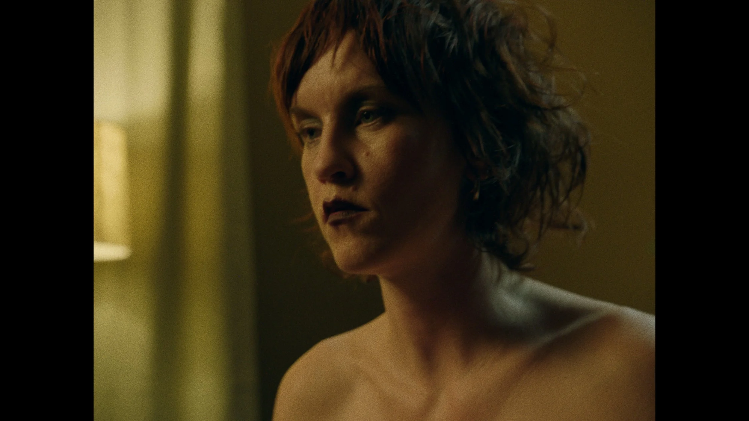 A woman with short, wavy reddish-brown hair looking down with a somber expression, in a dimly lit room.