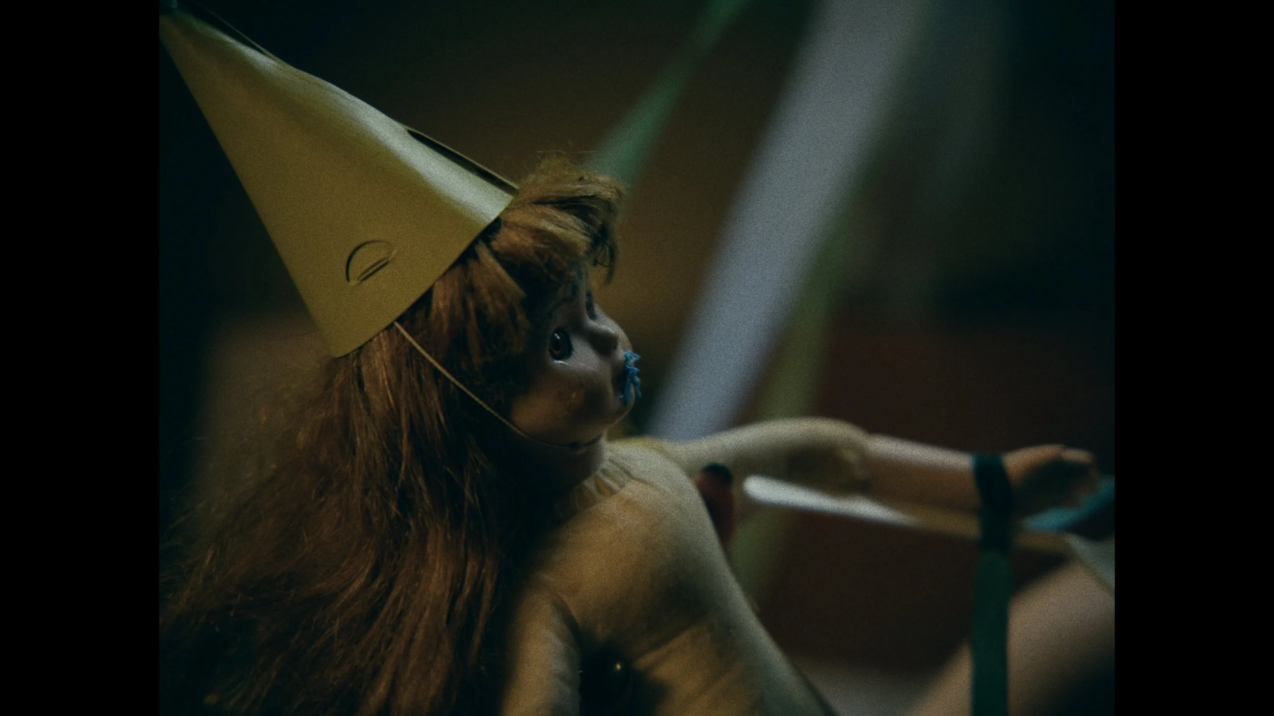 A doll with a human face, long brown hair, wearing a yellow cone-shaped party hat and a diaper, sitting with one arm extended, in a dimly lit environment.
