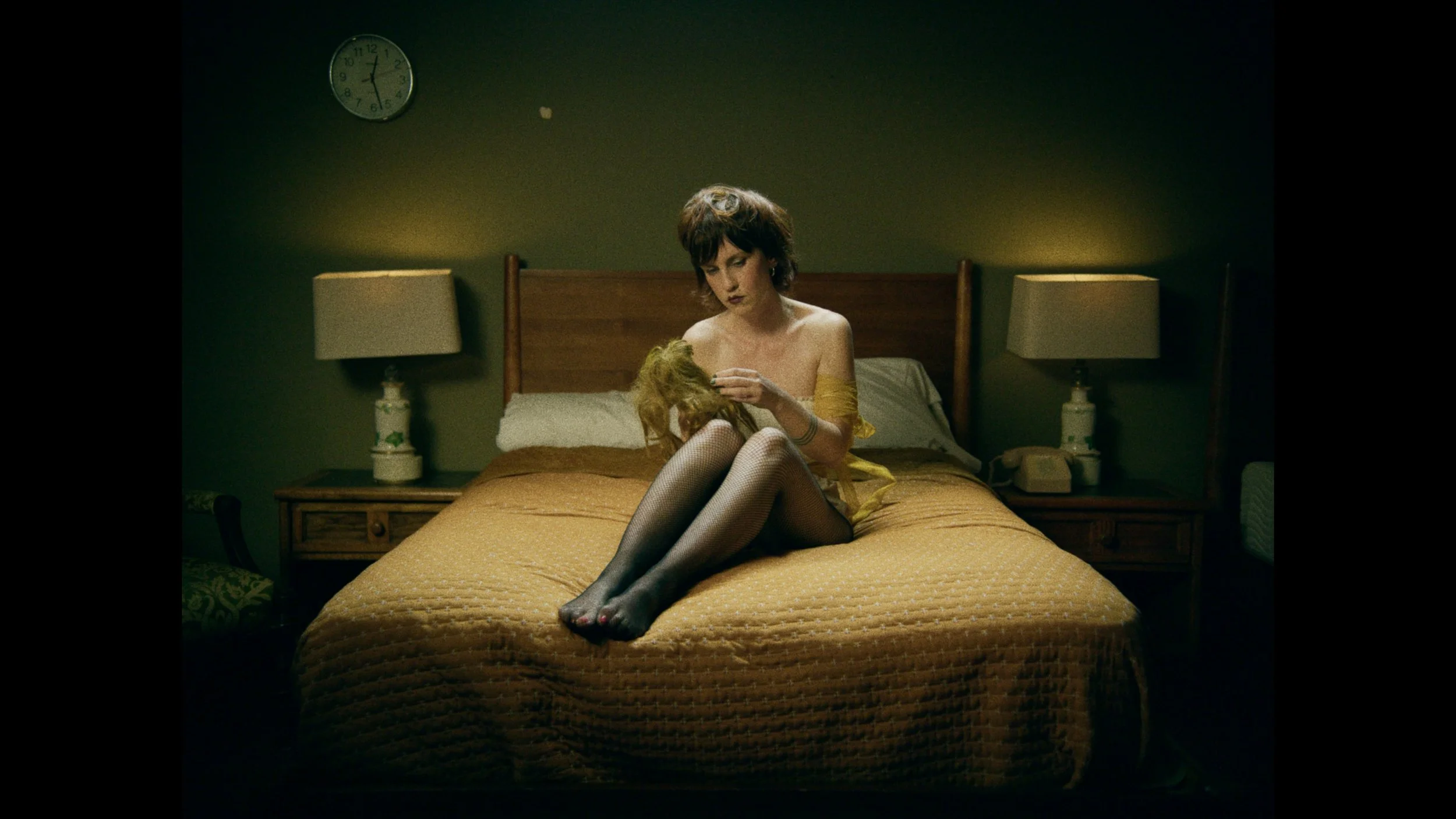 A woman with dark hair sitting on a bed, holding a blonde doll, in a dimly lit bedroom with a clock showing 12:23 on the wall and two table lamps on nightstands.