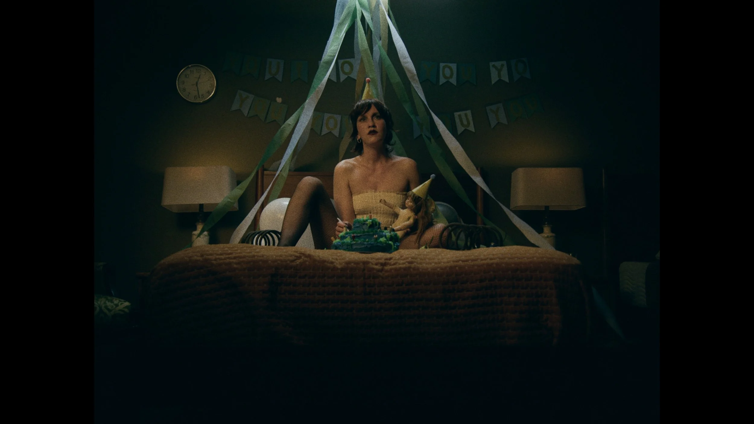 A woman sitting on a bed wearing a party hat with a birthday cake in front of her, surrounded by streamers and party decorations in a dimly lit room.