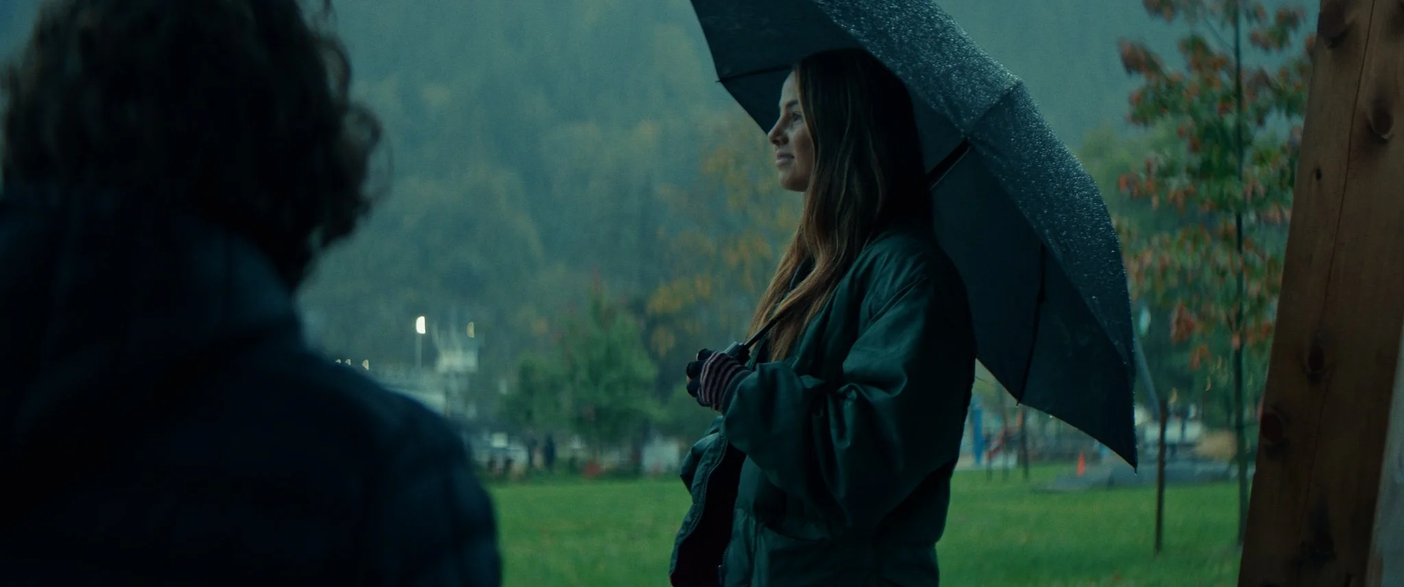 A woman standing outdoors in the rain holding an umbrella, engaging in conversation with a man, with trees and a park visible in the background.