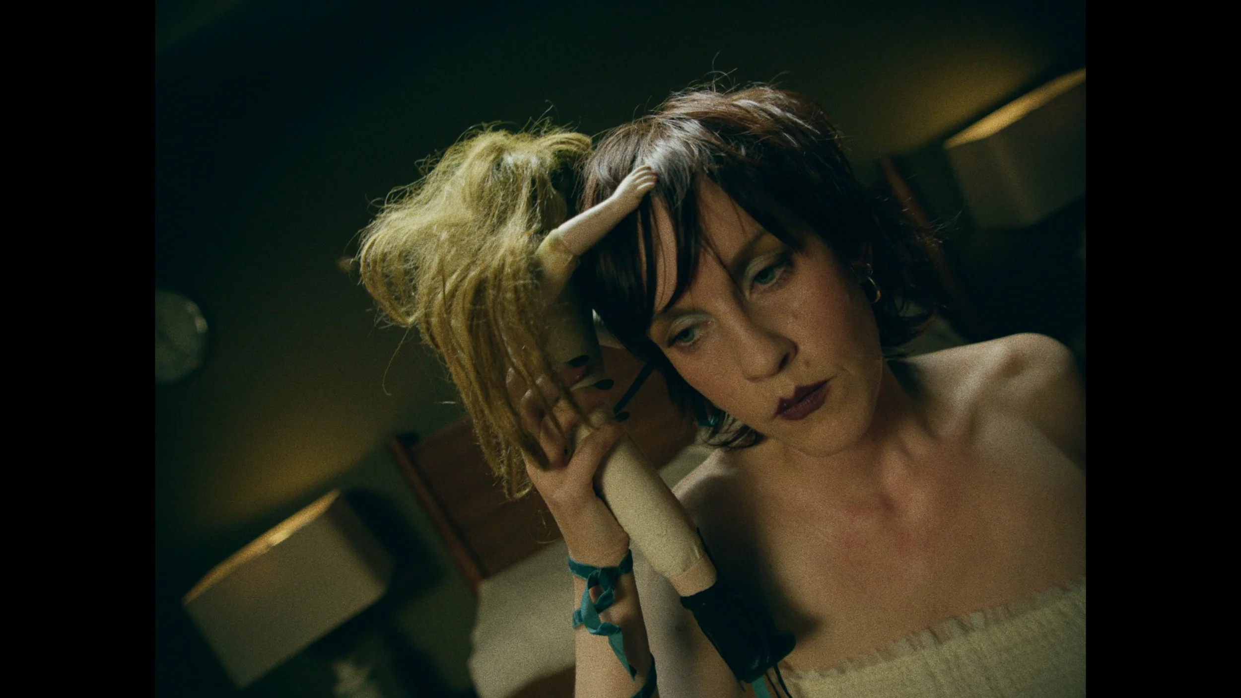 A woman with short dark hair and dark lipstick sits topless in a dimly lit room, holding a vintage telephone receiver to her ear with a pensive expression.