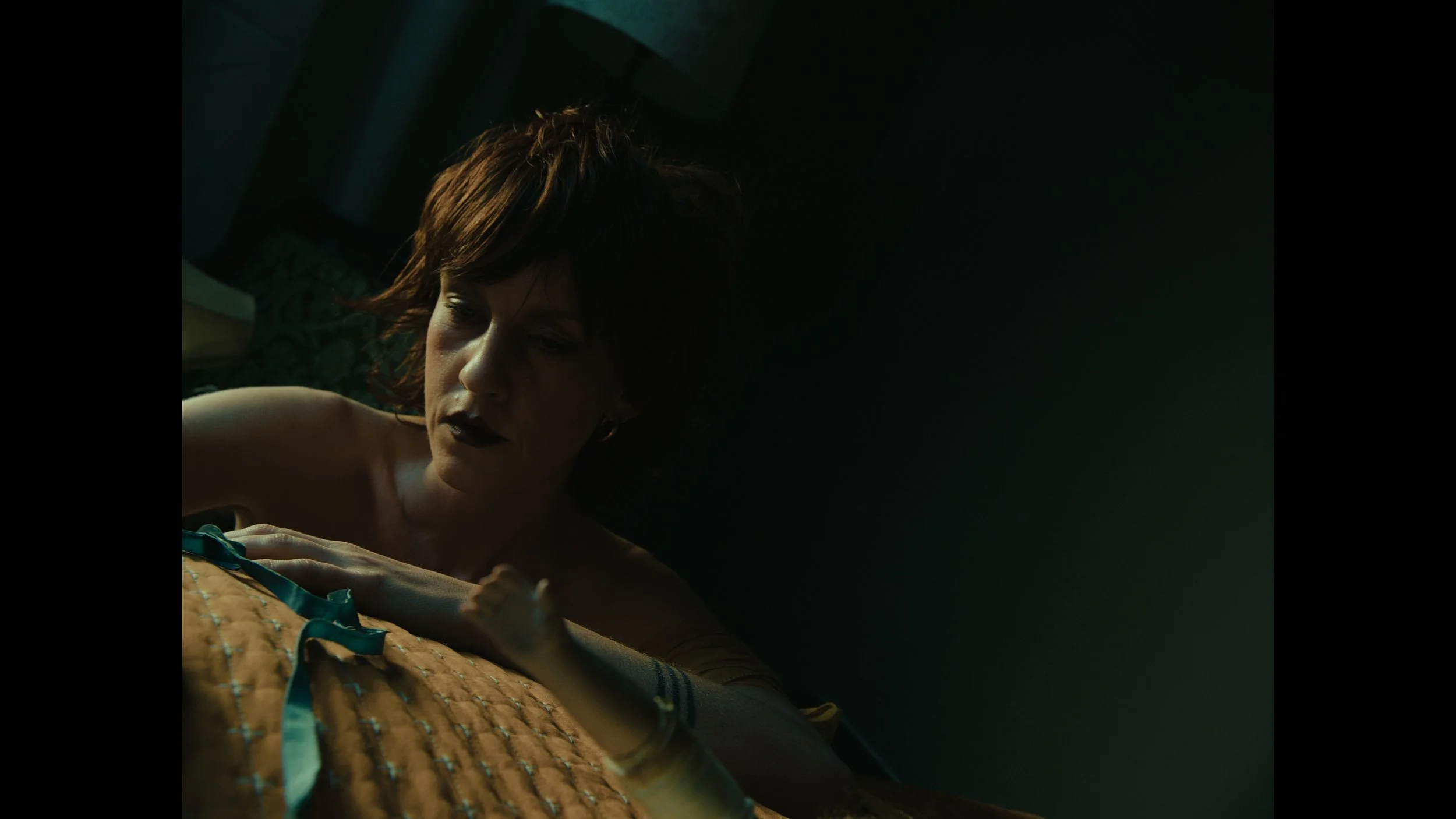 A woman with short, dark hair lying on a bed in a dimly lit room, staring at an object she is holding in her hand. The lighting and mood suggest a moody or intense scene.