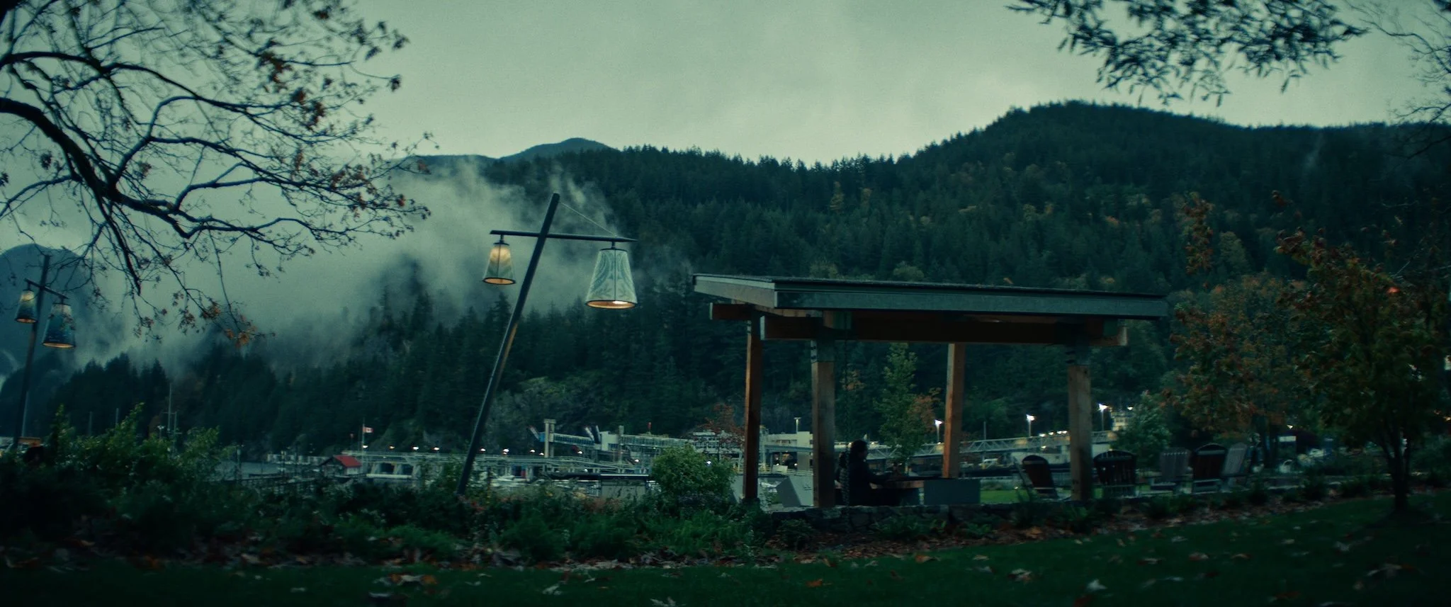 Outdoor scene with mountains, a cloudy sky, trees, and a marina with boats. There is a covered seating area with chairs, decorative street lamps, and a person sitting at a table.