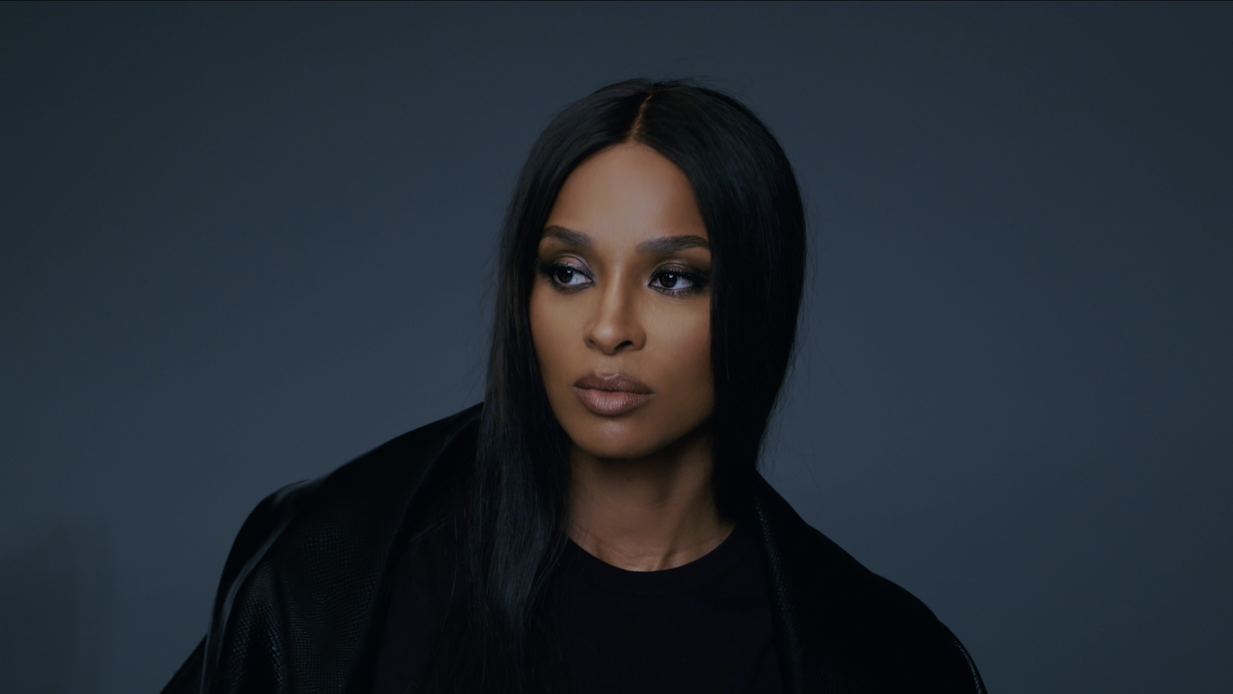 A woman with long black hair styled straight, wearing dark clothing, looking to her left against a dark gray background.