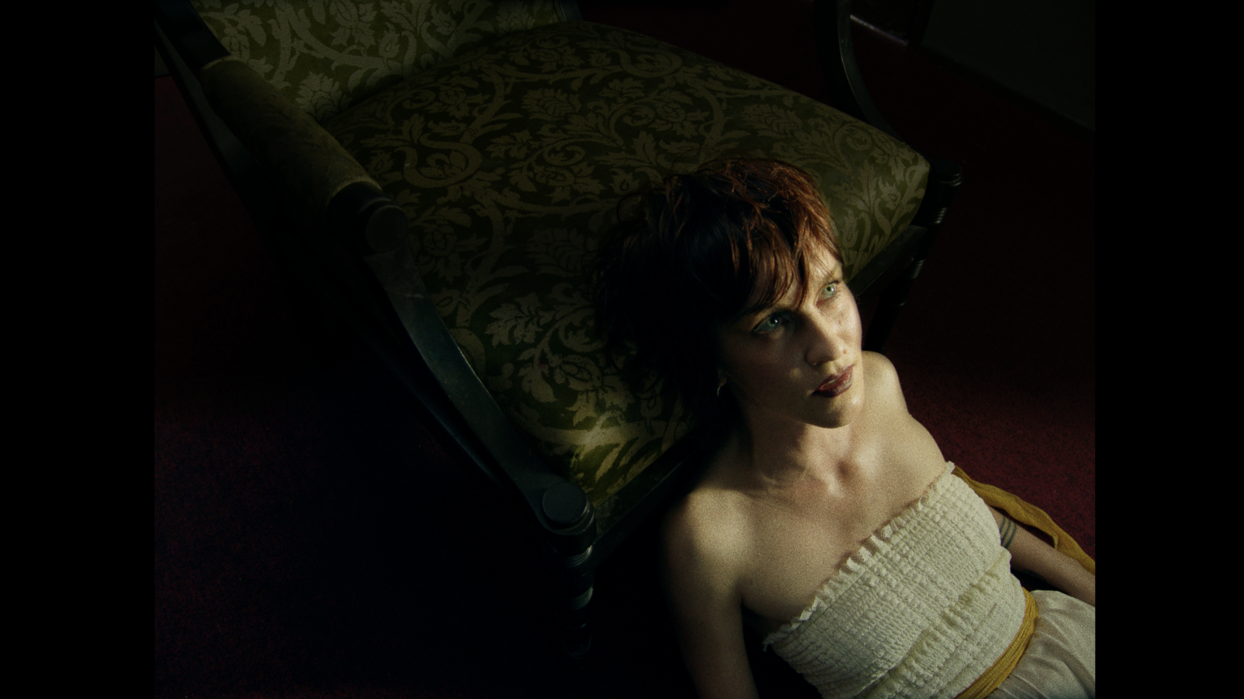 A woman with short reddish-brown hair and light skin gazes upward while sitting on the floor, leaning against a vintage-style wooden bench with green upholstery with light-colored floral pattern, illuminated by soft natural light.