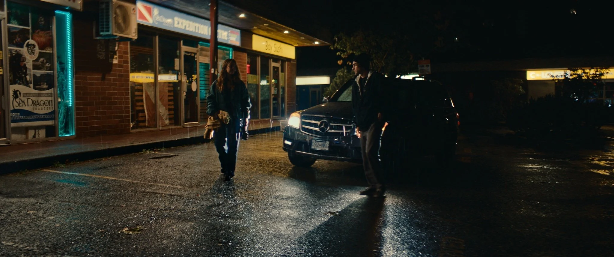 A man and woman standing outside a store on a rainy night, near a black SUV with its headlights on. The store has bright signs, a brick exterior, and illuminated windows, with rain-soaked pavement reflecting the lights.