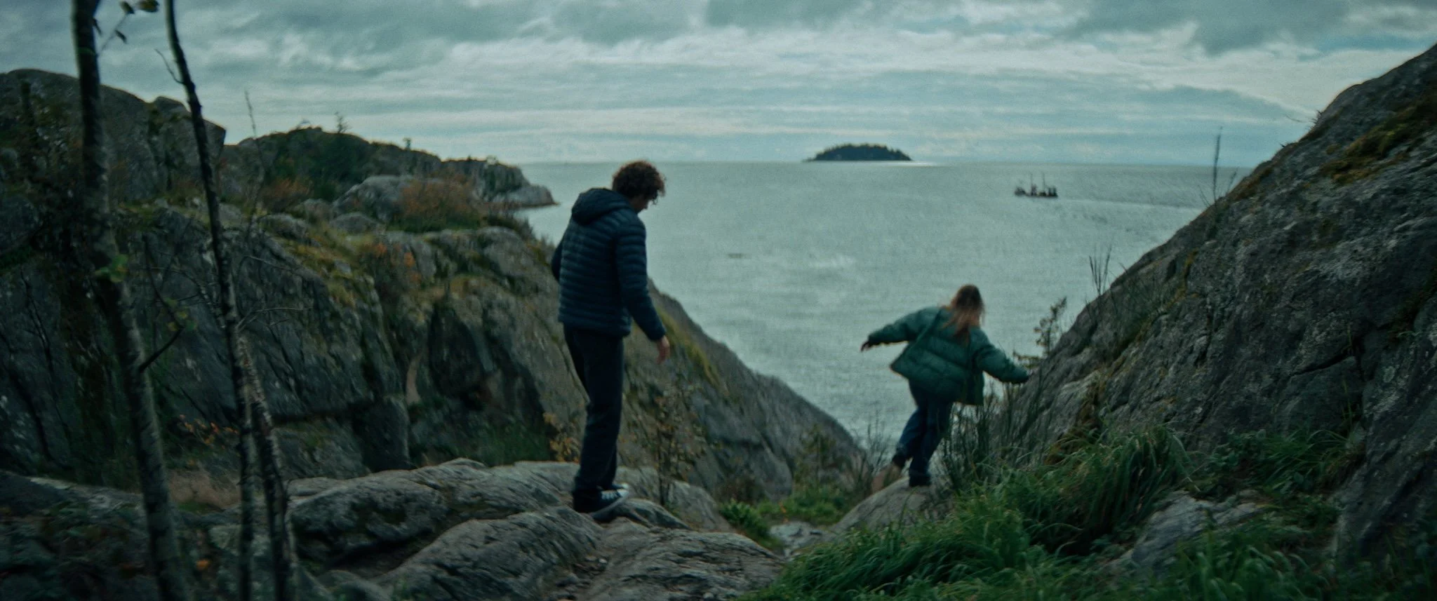 Two children, a boy and a girl, exploring rocky terrain near a body of water with an island in the distance, cloudy sky, and a boat.