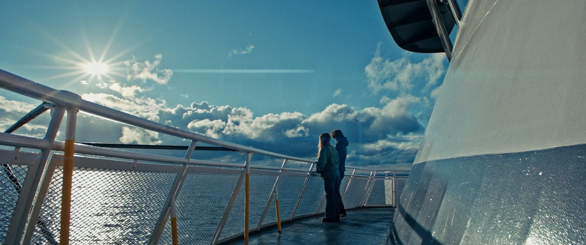 Two people standing on the deck of a ship, looking out at the ocean under a blue sky with clouds and the sun shining.